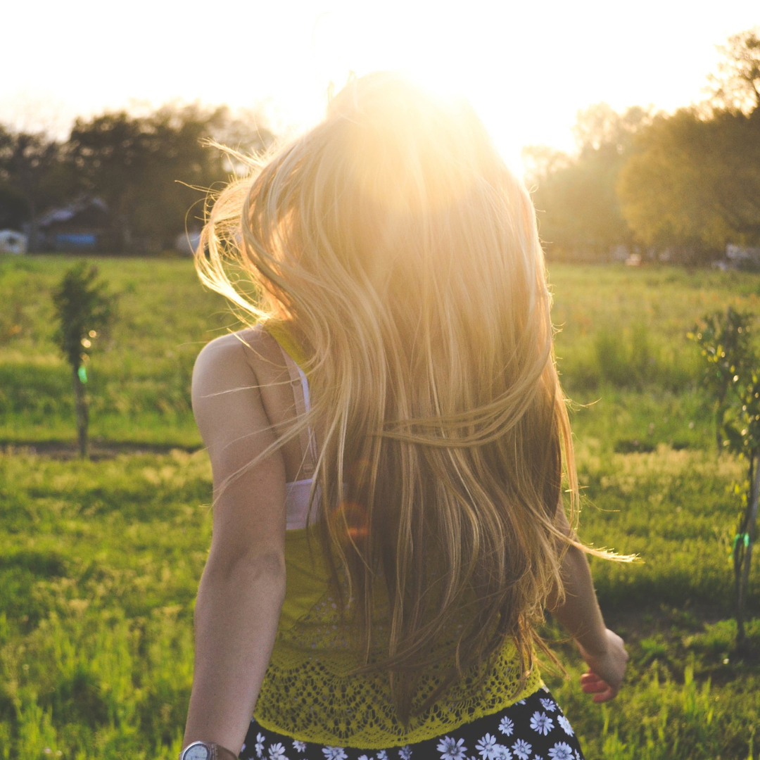 **Image description (alt text):** A person with long blonde hair is seen from behind walking through a green field at sunset, sunlight glowing around their head and hair, with trees in the distance.